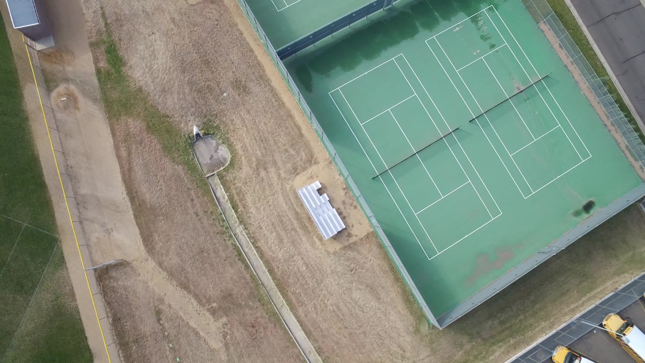 Aerial frame over Hidden Lake, Colorado showing two green tennis courts, a strip of dry ground, small white bleachers, grass fields, and no visible people.