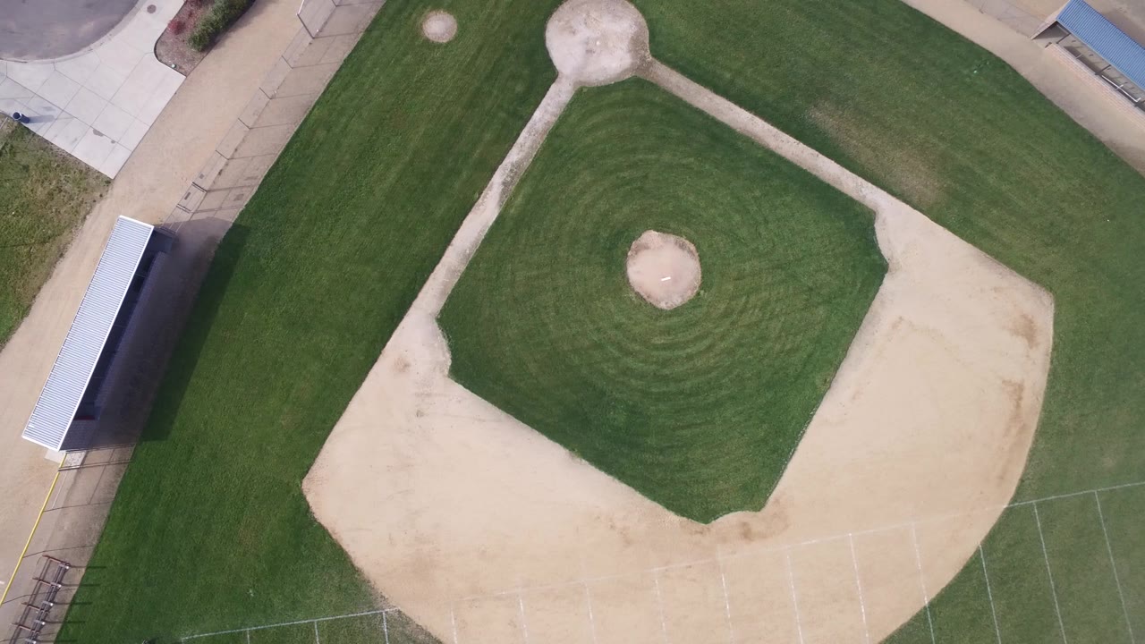 High overhead view in Hidden Lake, Colorado, showing the baseball infield centered in frame, pale dirt around home plate, a left-side shelter, paved paths, and no clearly visible people.
