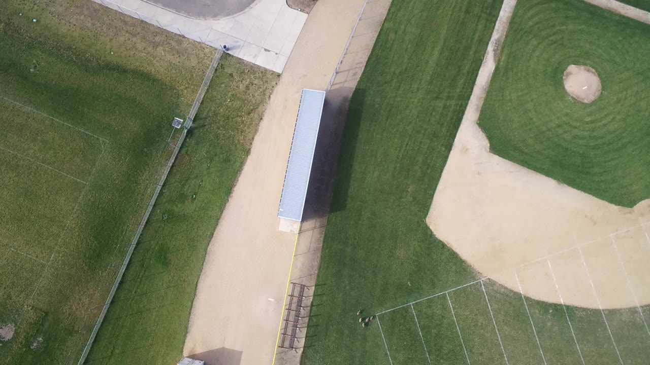 Second overhead frame in Hidden Lake, Colorado with the tan infield more centered, a long light-roofed shelter along the path, grass bands, and no visible people.