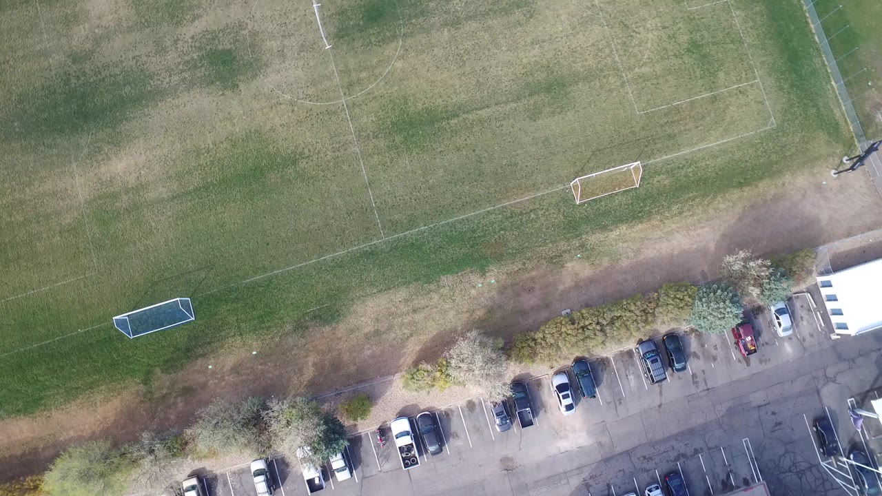 Overhead view of soccer fields, parked cars, trees, and building edge.