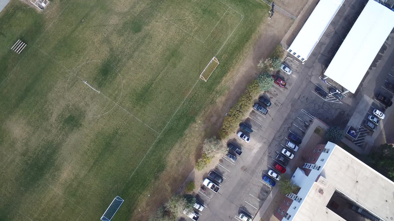 Overhead view of a green sports field beside a parking lot and flat-roofed buildings.