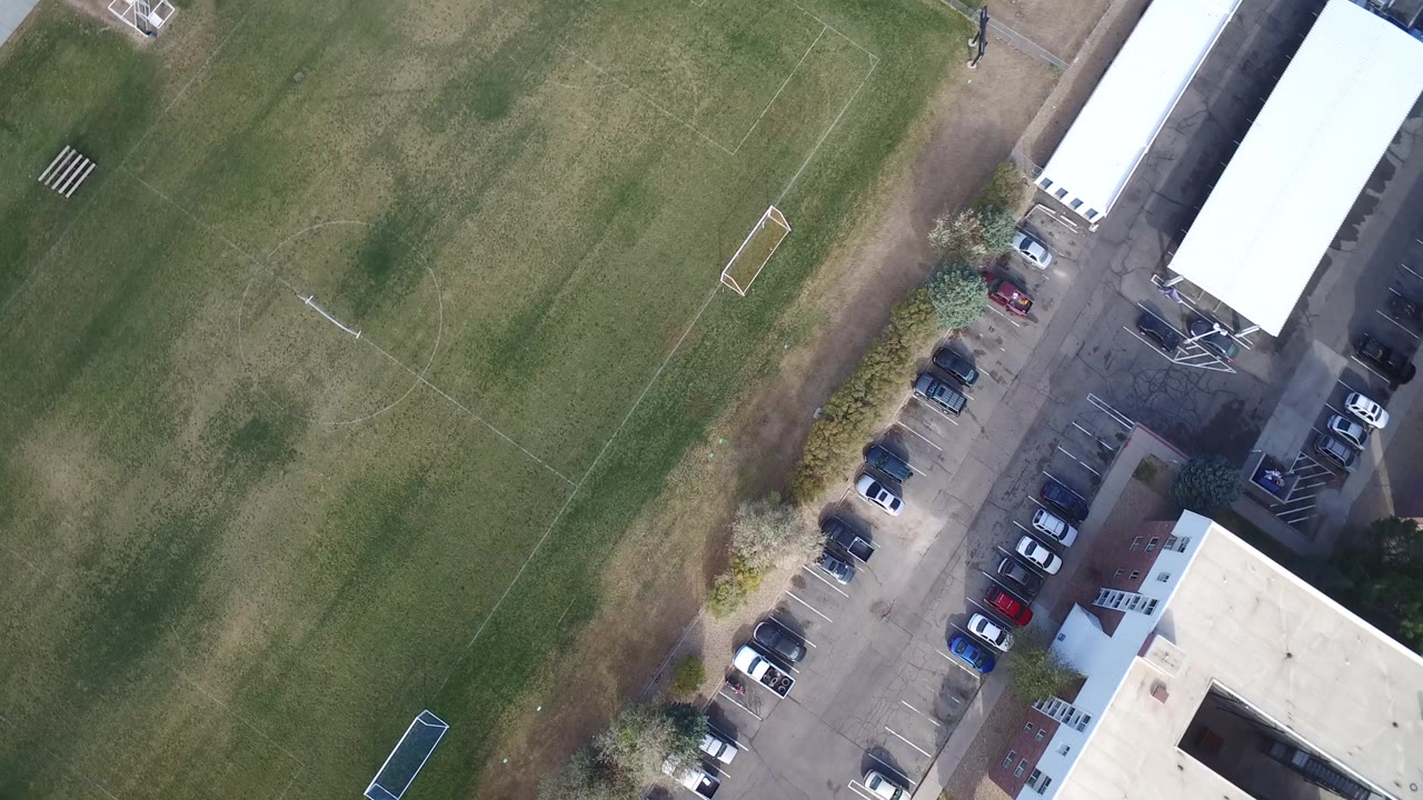 Second overhead frame in Hidden Lake, Colorado with the grassy pitch centered more fully, a row of parked cars along the lane, and white-roofed structures on the right, with no visible people.