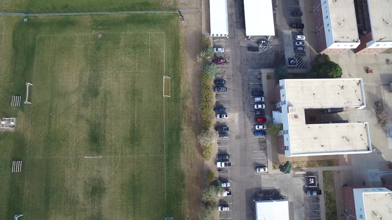 High overhead view in Hidden Lake, Colorado showing the left side of a marked soccer field, adjacent flat-roof buildings, a parking lane, parked cars, and no visible people.