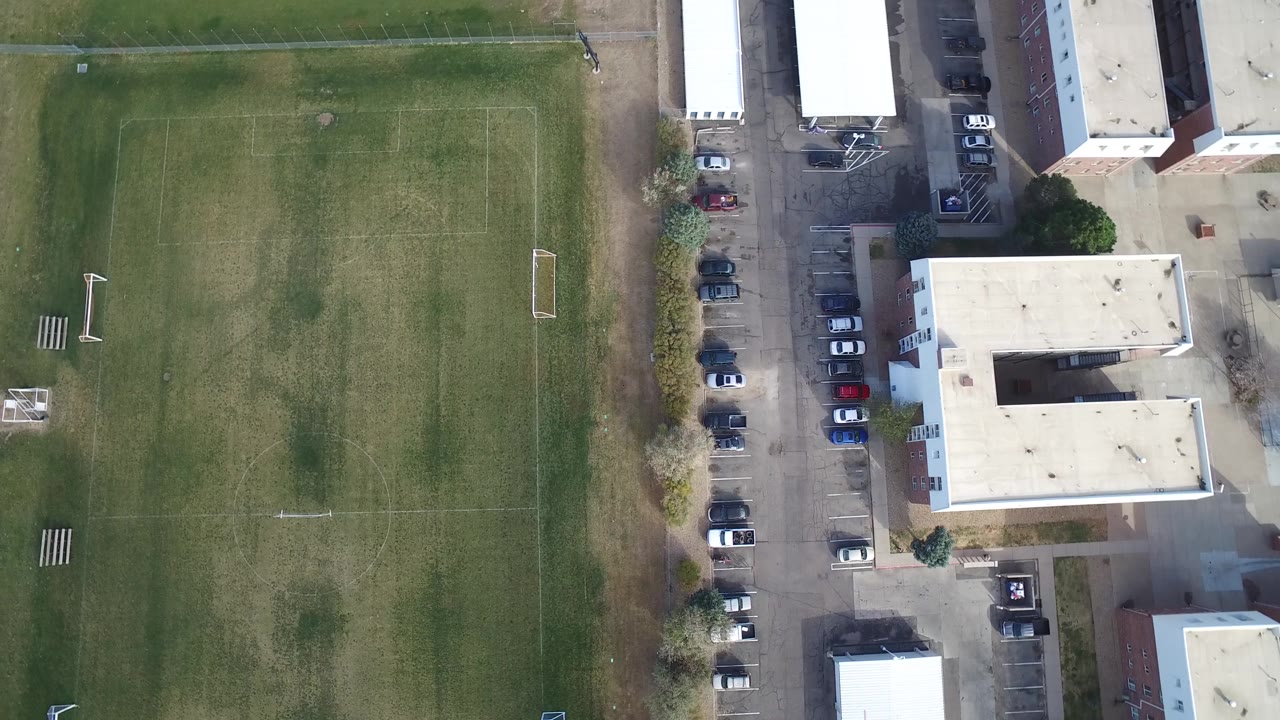 Overhead Hidden Lake, Colorado frame with the green soccer field centered left, pale-roof buildings to the right, parked cars along the drive, and no visible people.