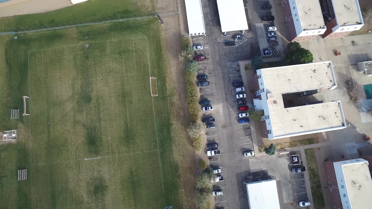 Top-down view of a soccer field, parking area, and flat-roof buildings.