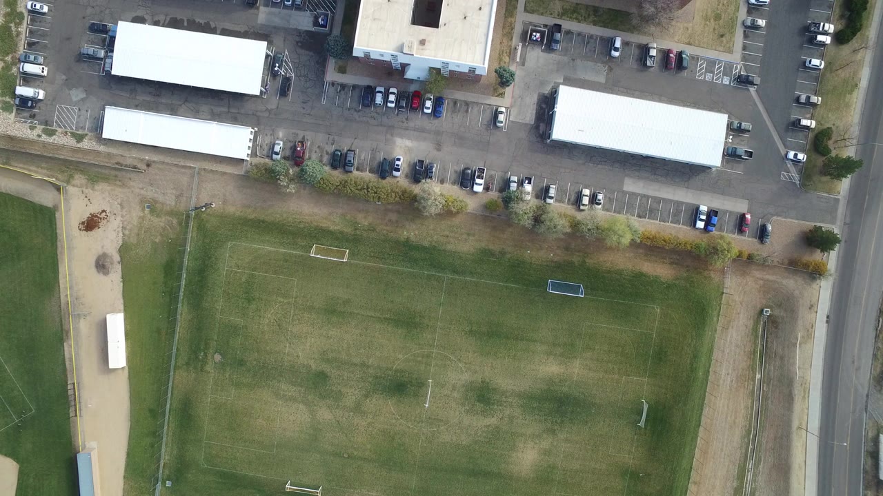 Overhead view of a grass sports field beside a parking lot and buildings.