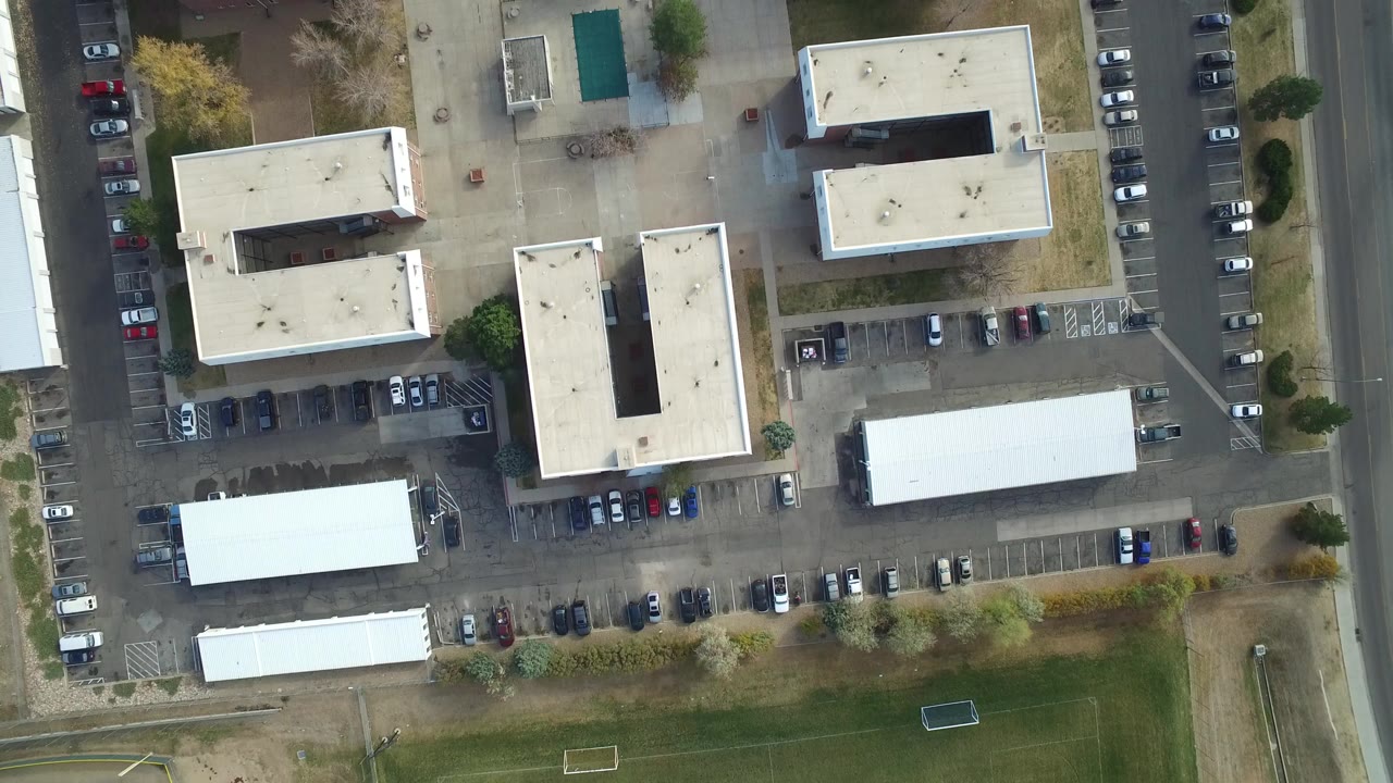 Top-down aerial view of low-rise buildings, parking lots, road, and grass field.