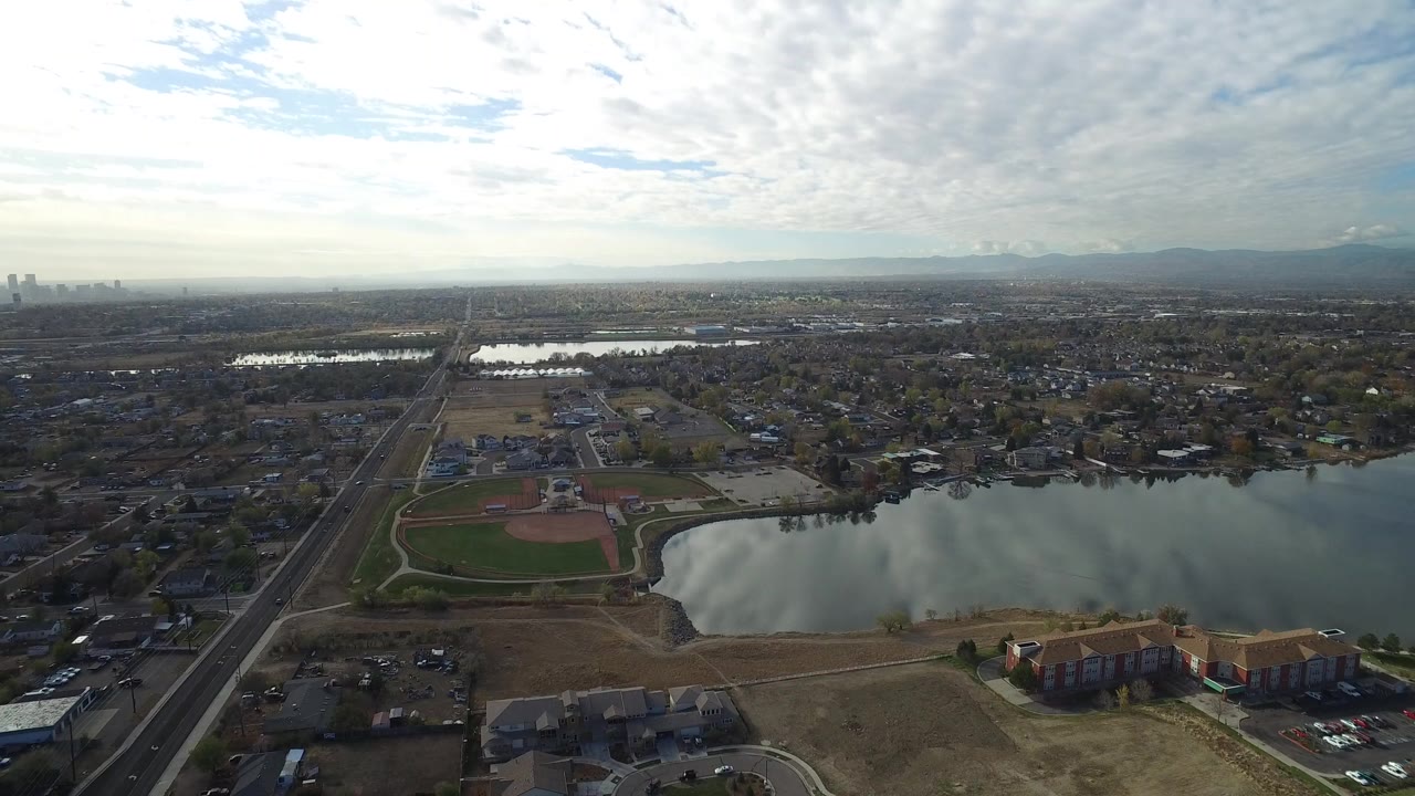 Aerial view of a lake, ball fields, roads, homes, and distant skyline under clouds.