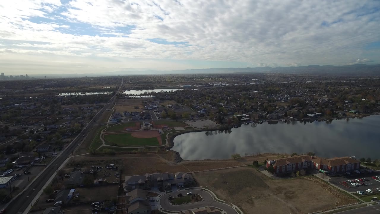 Opening aerial frame over Hidden Lake, Colorado, with a reflective lake beside apartments, a baseball field at left, suburban streets beyond, distant mountains, and no visible people.