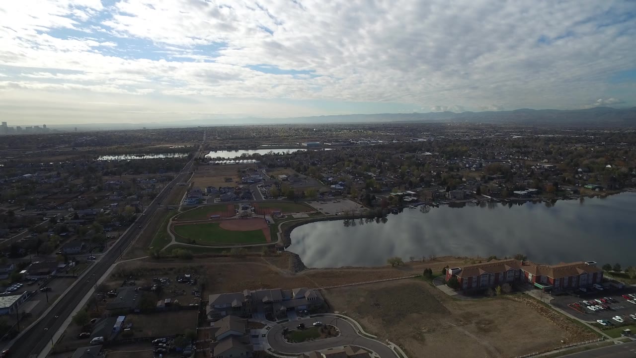 Early aerial view of Hidden Lake, Colorado, showing the lake centered between apartments in the foreground and a spread of homes beneath bright layered clouds, with no visible people.
