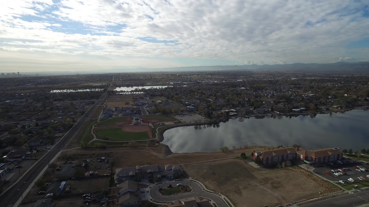 Wide aerial over a lake, ball field, apartment complex, streets, and distant mountain ridge.