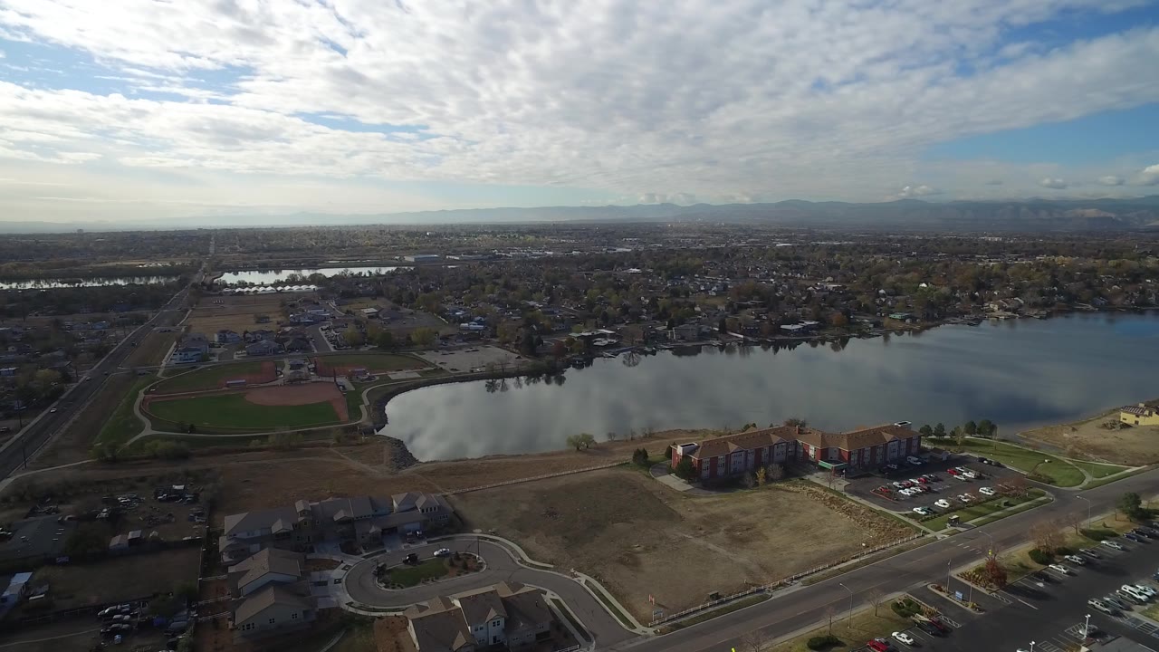 Aerial view of a lake beside sports fields, roads, apartments, and distant neighborhoods.