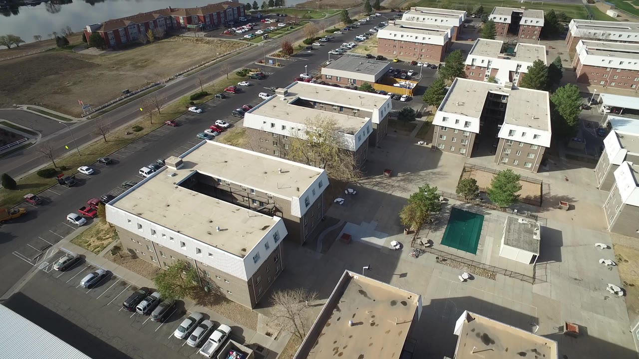 Early overhead view in Hidden Lake, Colorado, centered on apartment rooftops, a paved courtyard, parking rows, trees, and the road beside the complex with no visible people.