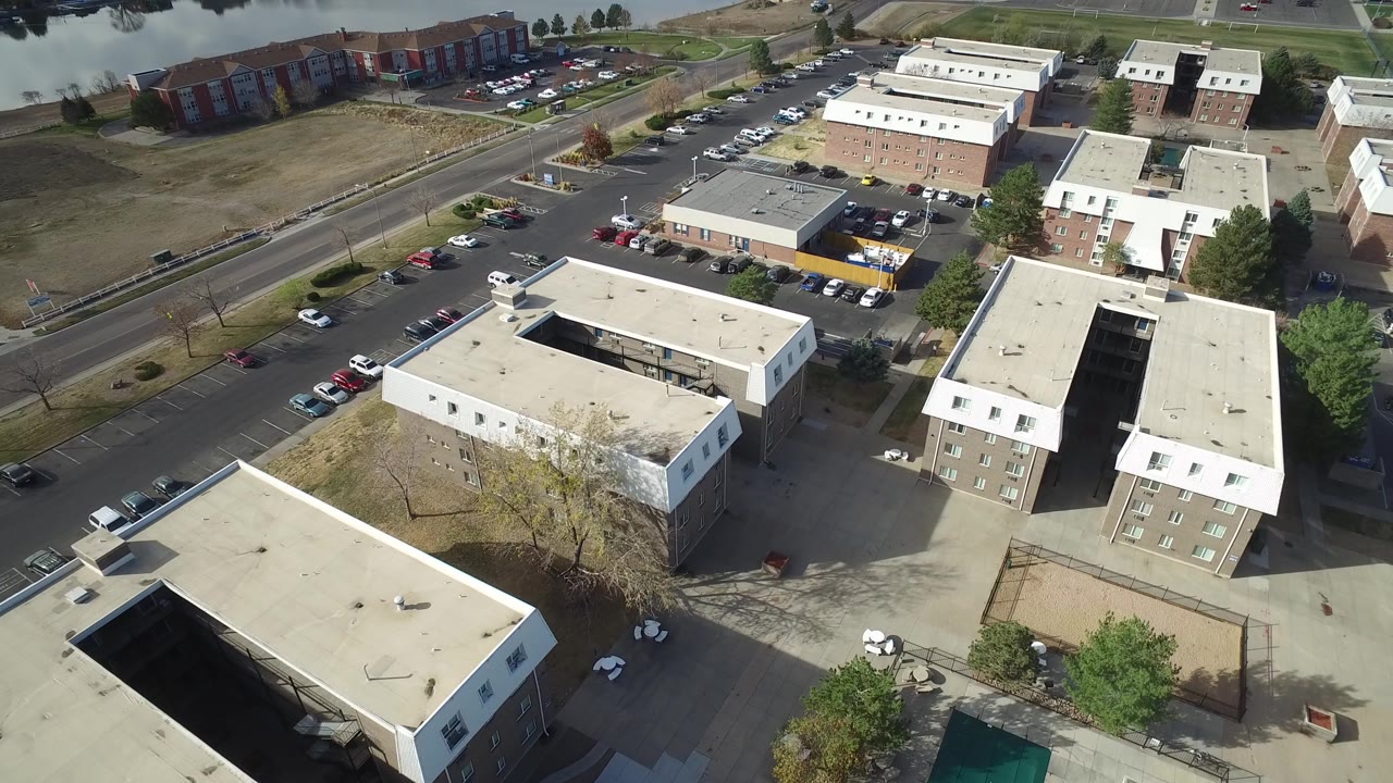 Early aerial frame in Hidden Lake, Colorado showing the camera over flat apartment roofs, a central parking lot, roadside cars, open grass, and a narrow band of water, with no visible people.