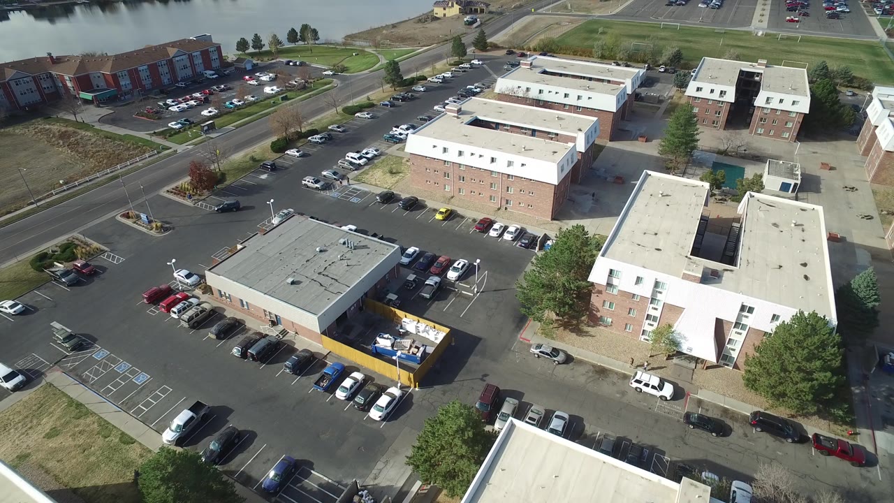 Aerial frame over Hidden Lake, Colorado with brick buildings centered, rows of parked cars below, a road crossing the upper frame, and no visible people.