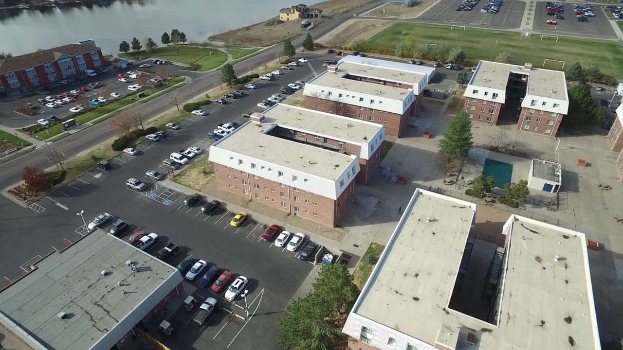 Aerial view of brick buildings, flat roofs, parked cars, roads, and water beyond the lot.