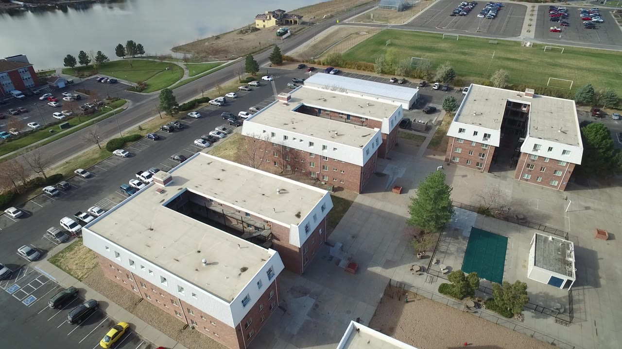 High aerial view in Hidden Lake, Colorado showing flat-roofed brick buildings, parked cars, a road, grass fields, and water along the upper edge, with no visible people.
