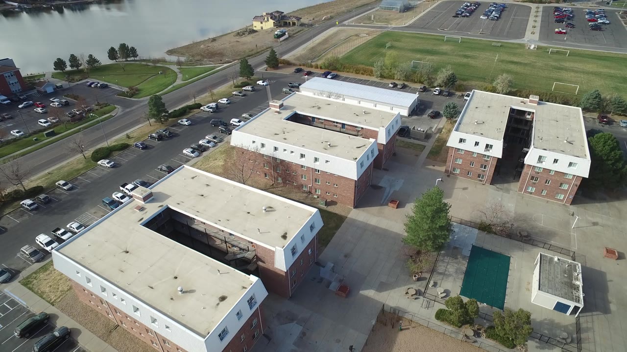 Drone-like overhead view of buildings, a parking lot, a road, sports fields, and water.