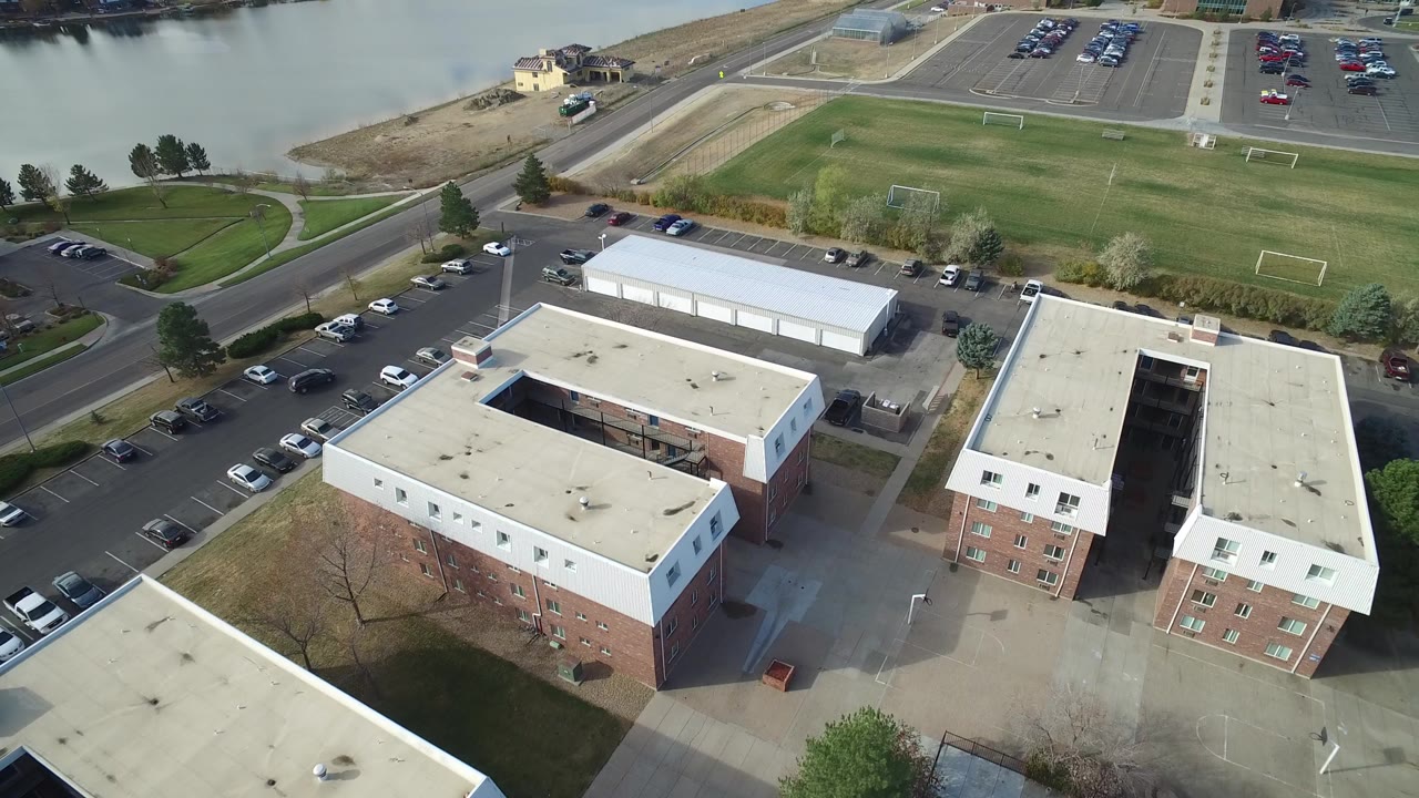 Aerial view of flat-roof buildings, parking lots, fields, roads, and lake water.