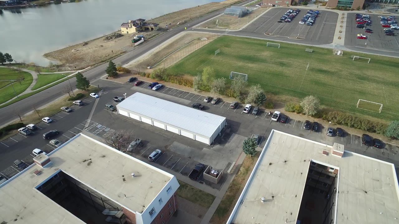 Opening aerial frame over Hidden Lake, Colorado, with a green soccer field, white goals, parked cars, flat-roofed buildings, roads, shoreline, pale water, and no visible people.