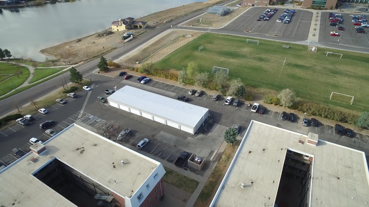 Aerial frame in Hidden Lake, Colorado, showing more of the soccer field center with parking rows above it, low buildings below it, the lake edge in the background, and no visible people.