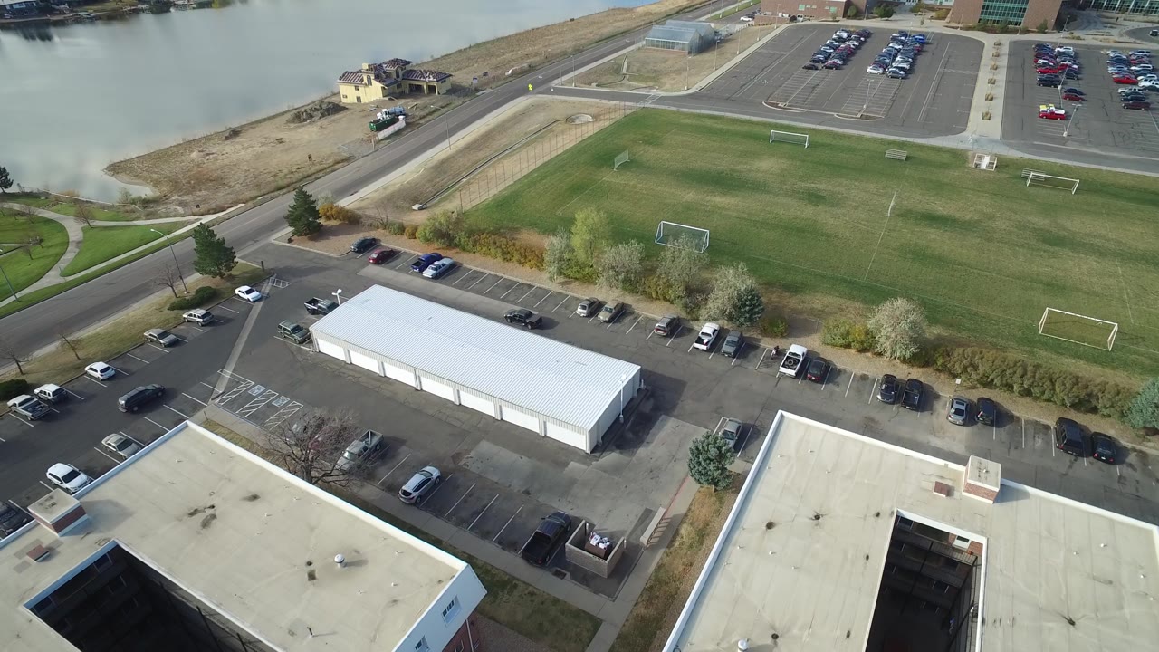 Overhead view of a soccer field, parked cars, roads, buildings, and shoreline.