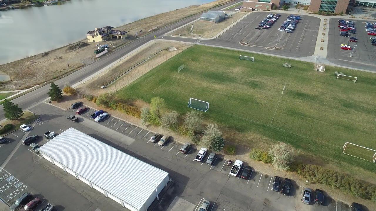 Aerial frame over Hidden Lake, Colorado showing a broad green field, a white-roofed building, parked cars, a road, and water at the upper left, with no visible people.