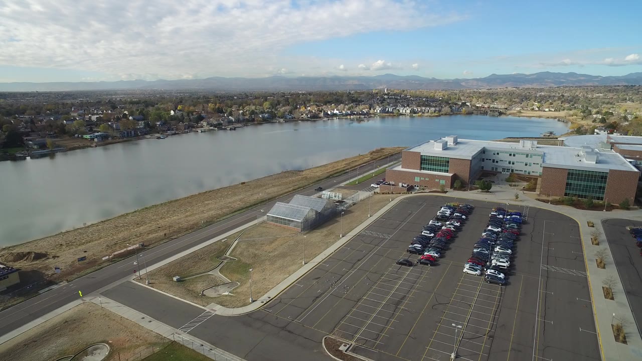 Opening aerial frame over Hidden Lake, Colorado showing a modern building beside the lake, a parking lot in the foreground, distant homes, mountains, blue sky, and no visible people.