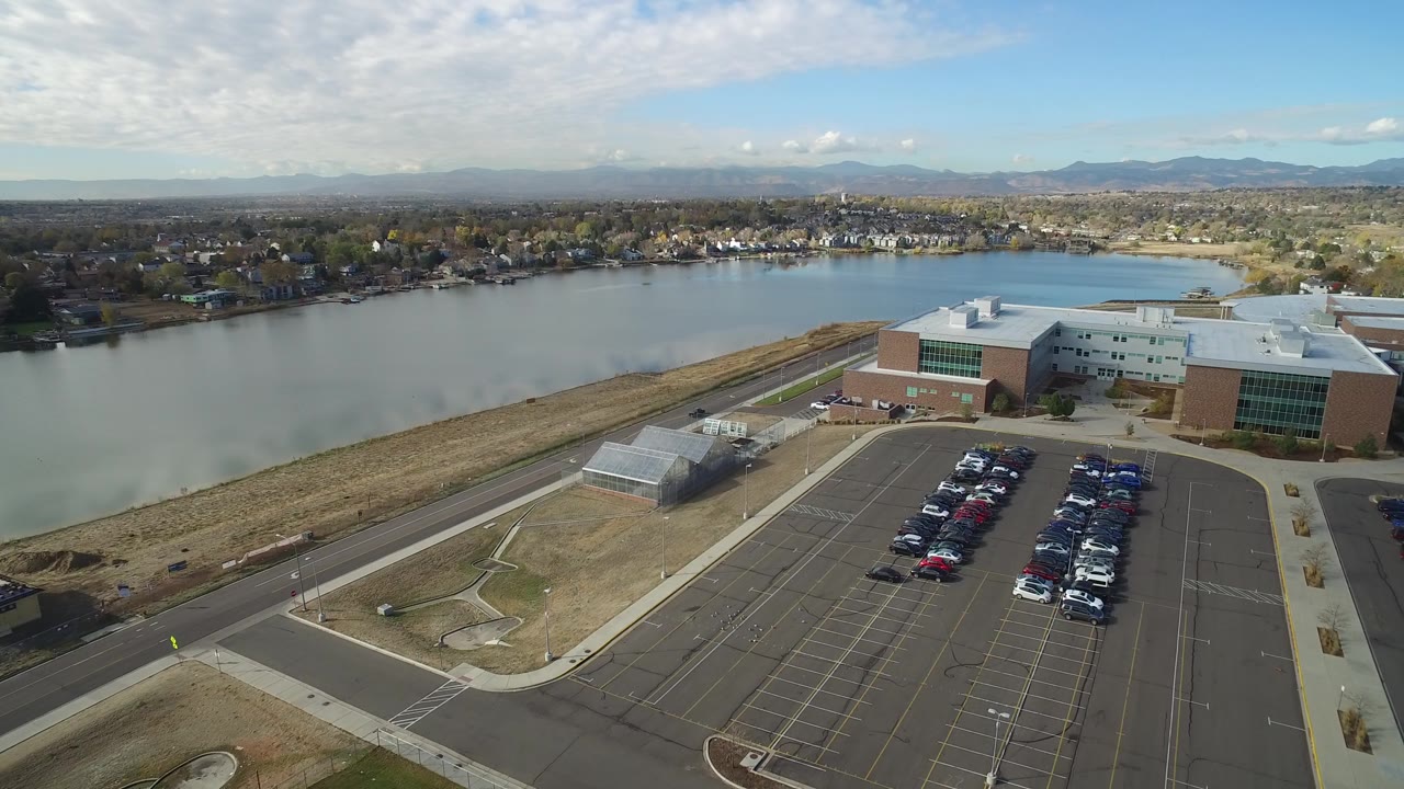 Aerial frame from Hidden Lake, Colorado with the lake stretching behind a modern brick-and-glass building, rows of parked cars, distant homes, mountains, and no visible people.