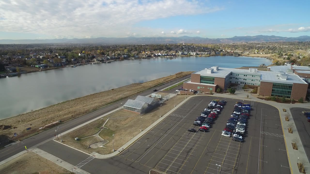 Drone view of a lake, shoreline homes, building roof, and parking lot.