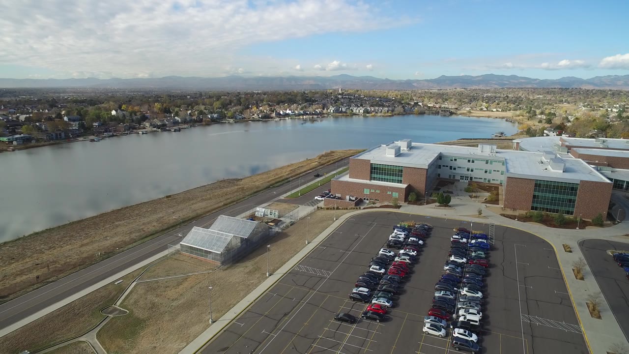 Aerial view of a lakeside brick building, parking lot, road, lake, houses, and mountains.