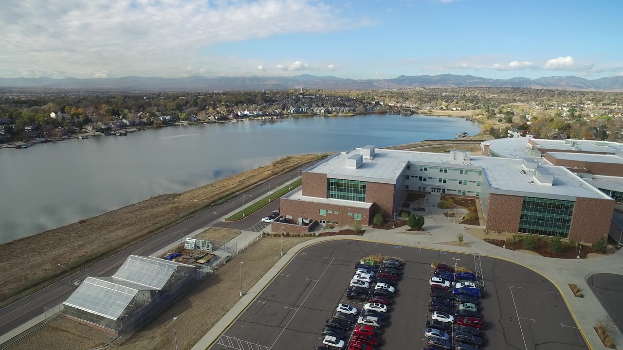 At 0:01, the Hidden Lake aerial angle keeps the building centered with the parking lot below, lake behind, residential shoreline across the water, and no visible people.
