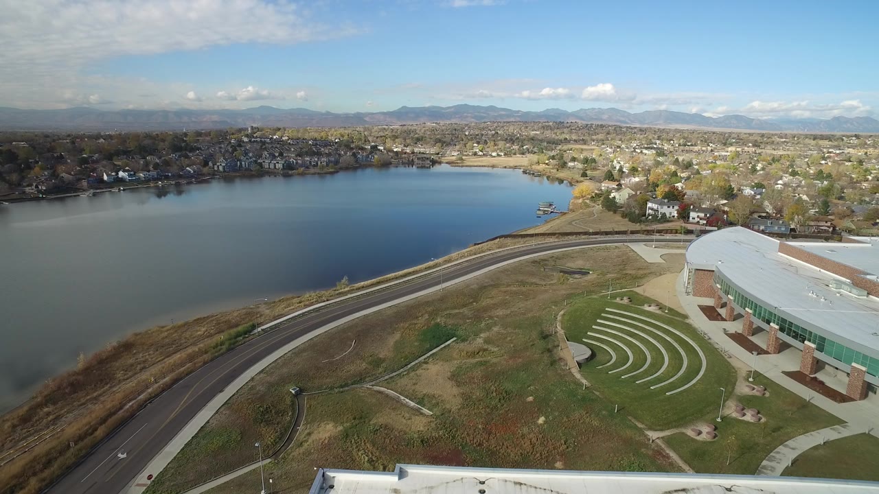 Aerial view of lake, road, modern building, homes, and distant mountains.