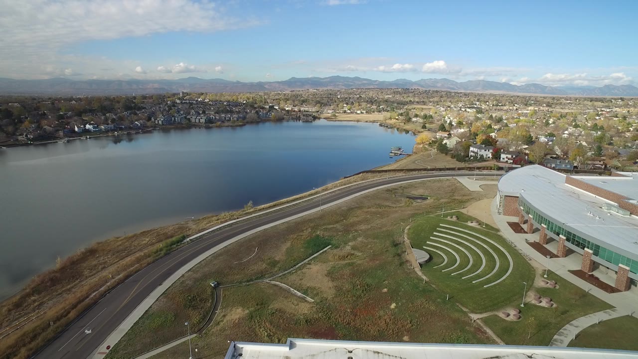 Hidden Lake, Colorado appears from above with the lake filling the left side, the road arcing through dry grass, the curved-roof building at lower right, homes across the water, and no visible people.