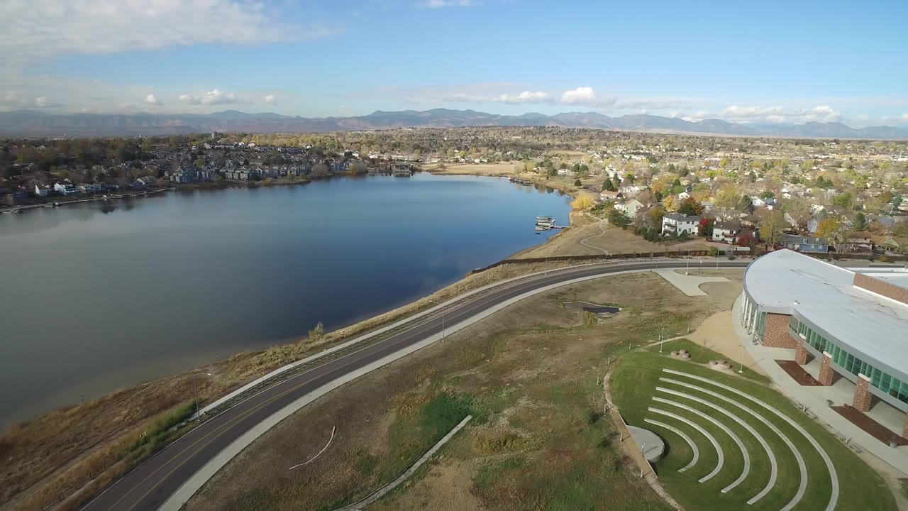Aerial view of a lake, curving road, modern building, homes, and distant mountains.