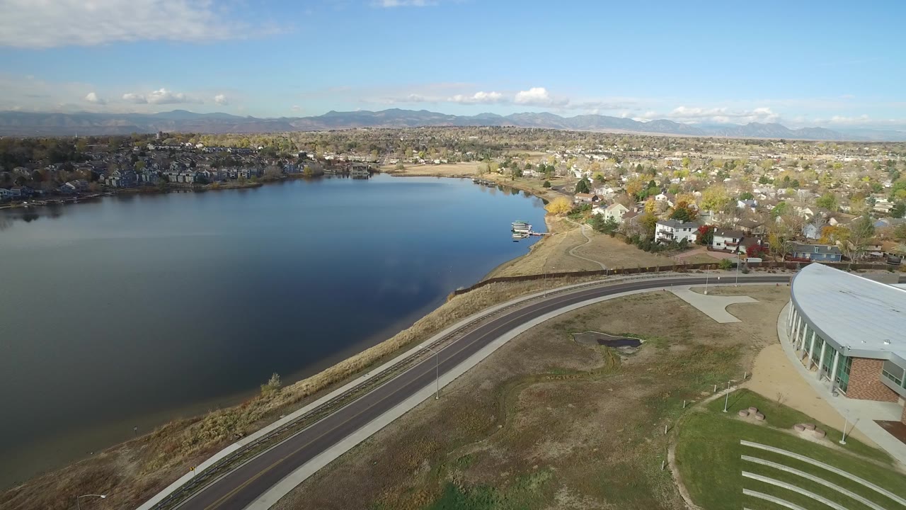 Aerial view of a lake, curved road, homes, trees, and distant mountains.