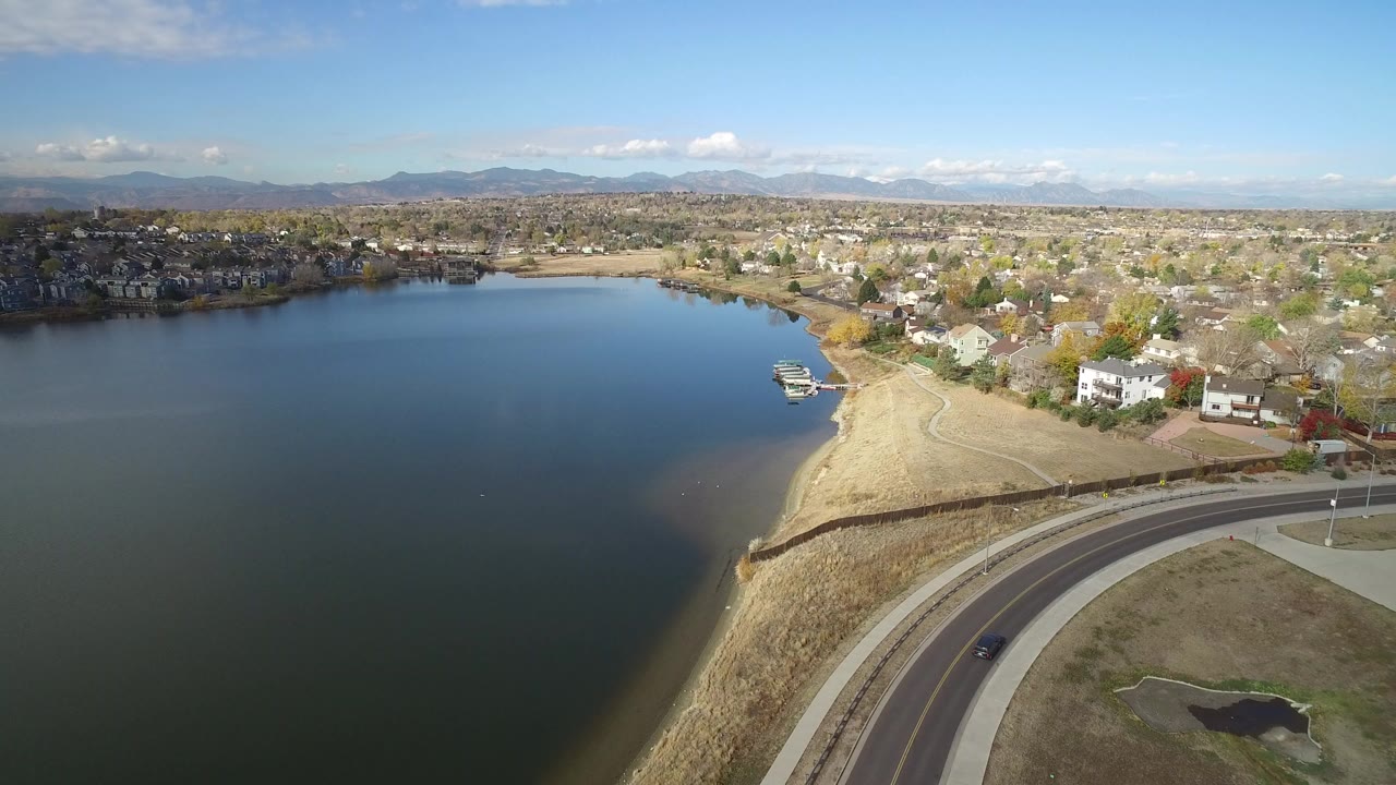 Opening aerial frame over Hidden Lake, Colorado, with dark blue water on the left, a curved lakeside road below, homes and trees beyond the shore, distant mountains, and no visible people.
