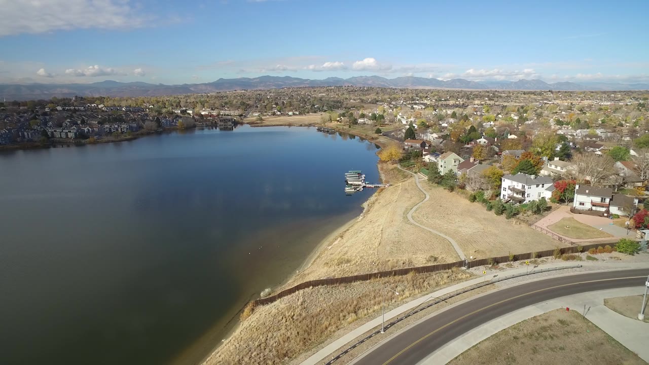 Wide drone-style view of Hidden Lake, Colorado with a walking path tracing the tan shore, docks near the water, houses along the right side, mountains on the horizon, and no visible people.