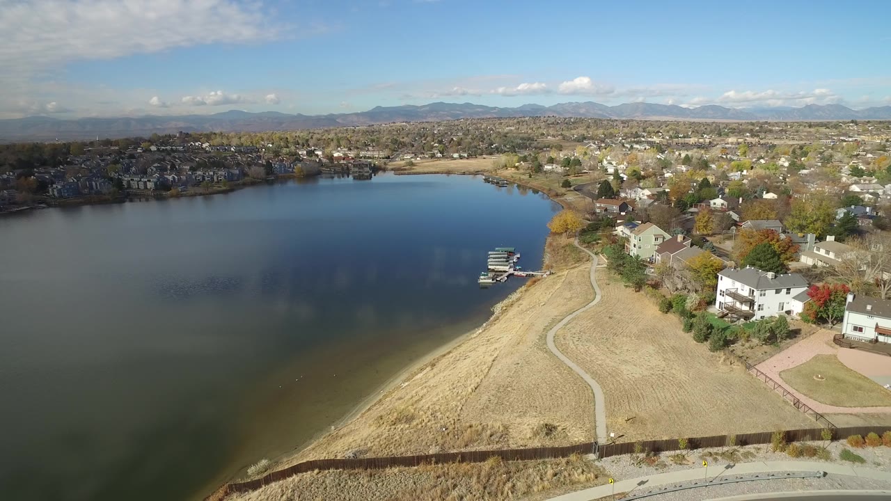Aerial view of lake, shoreline path, homes, dock, and distant mountains.