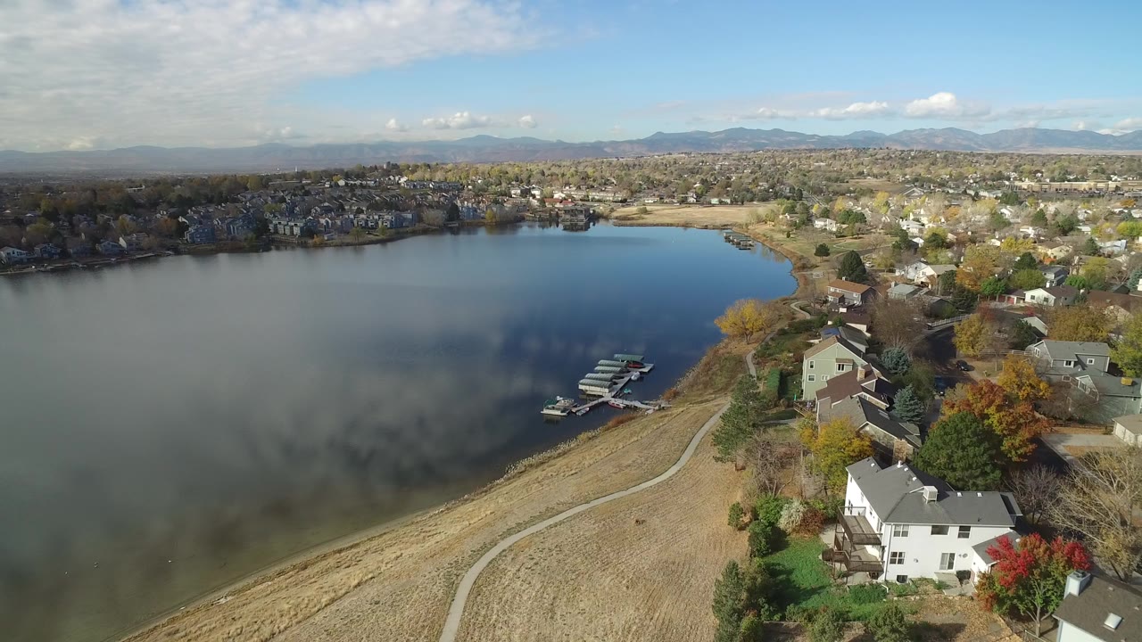 Aerial view over Hidden Lake, Colorado with open water on the left, a curved shoreline path, lakeside houses, docks, distant neighborhoods, mountains, and no visible people.