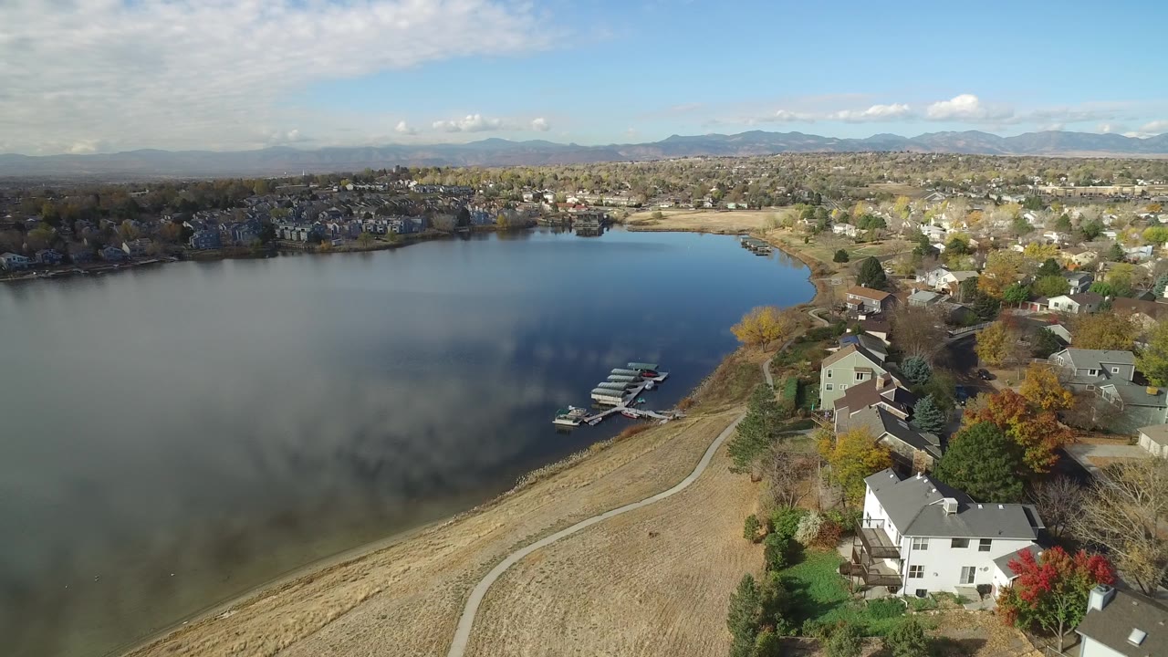 Aerial frame of Hidden Lake, Colorado with the dock cluster near the shoreline, homes along the lower right, a residential area across the water, mountains, and no visible people.