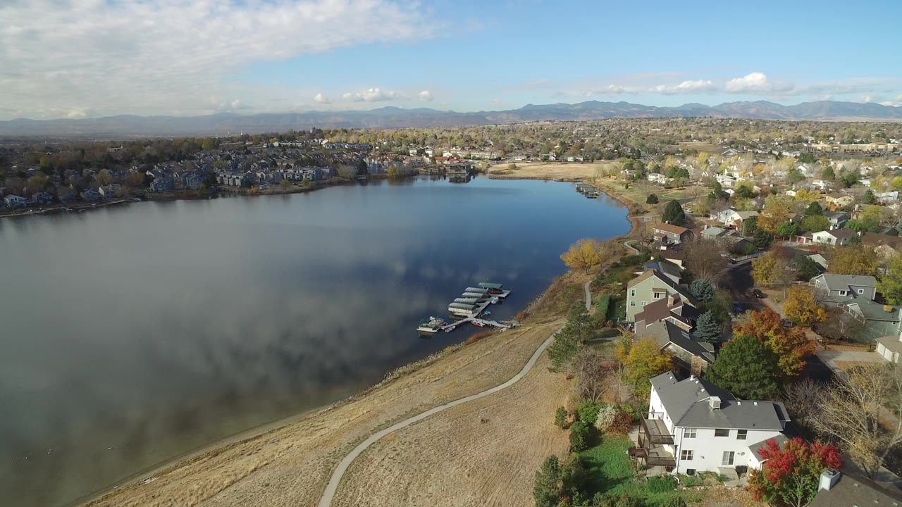 Drone view of calm lake water, shoreline homes, docks, and distant mountains.