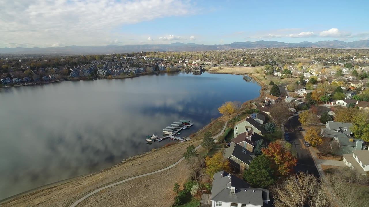 Drone-style aerial over Hidden Lake, Colorado, showing more of the shoreline docks, a green-roofed house, a curved path beside the water, distant homes, mountains, and no visible people.