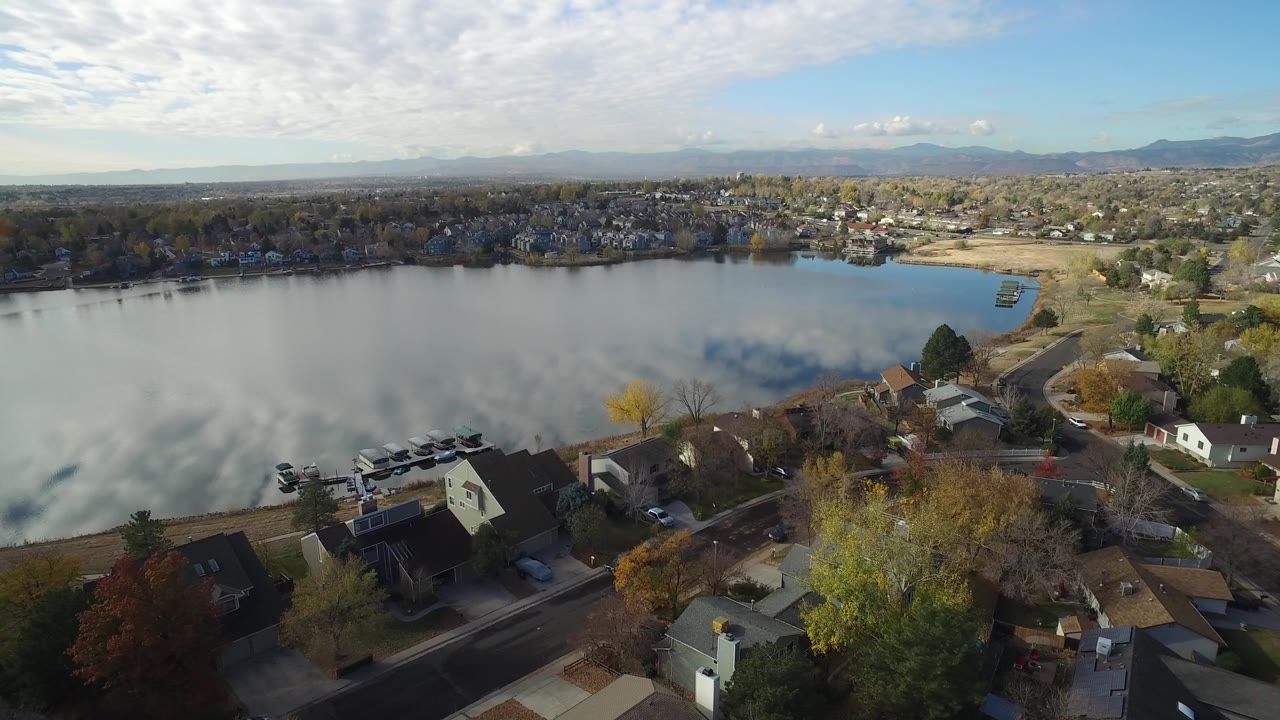 Aerial view over lakeside homes, a reflective lake, trees, and distant mountains.