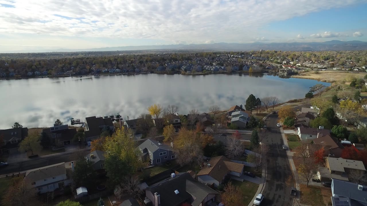 Opening aerial frame over Hidden Lake, Colorado, with residential rooftops in the foreground, still water across the middle, distant hills, and no visible people.