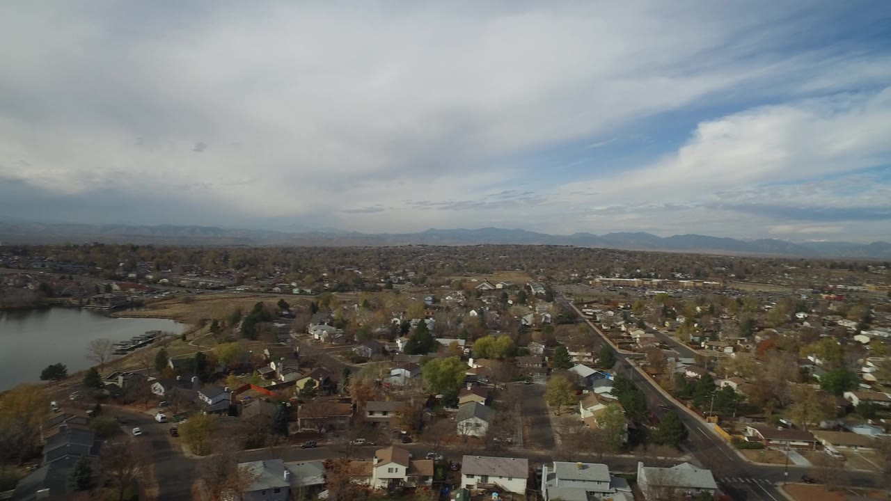 At 0:00, an aerial view of Hidden Lake, Colorado shows residential blocks, a lake edge on the left, distant mountains, and a heavy cloud band with no visible people.