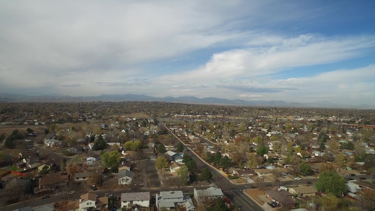 Aerial neighborhood panorama with roads, homes, trees, distant mountains, and layered clouds.