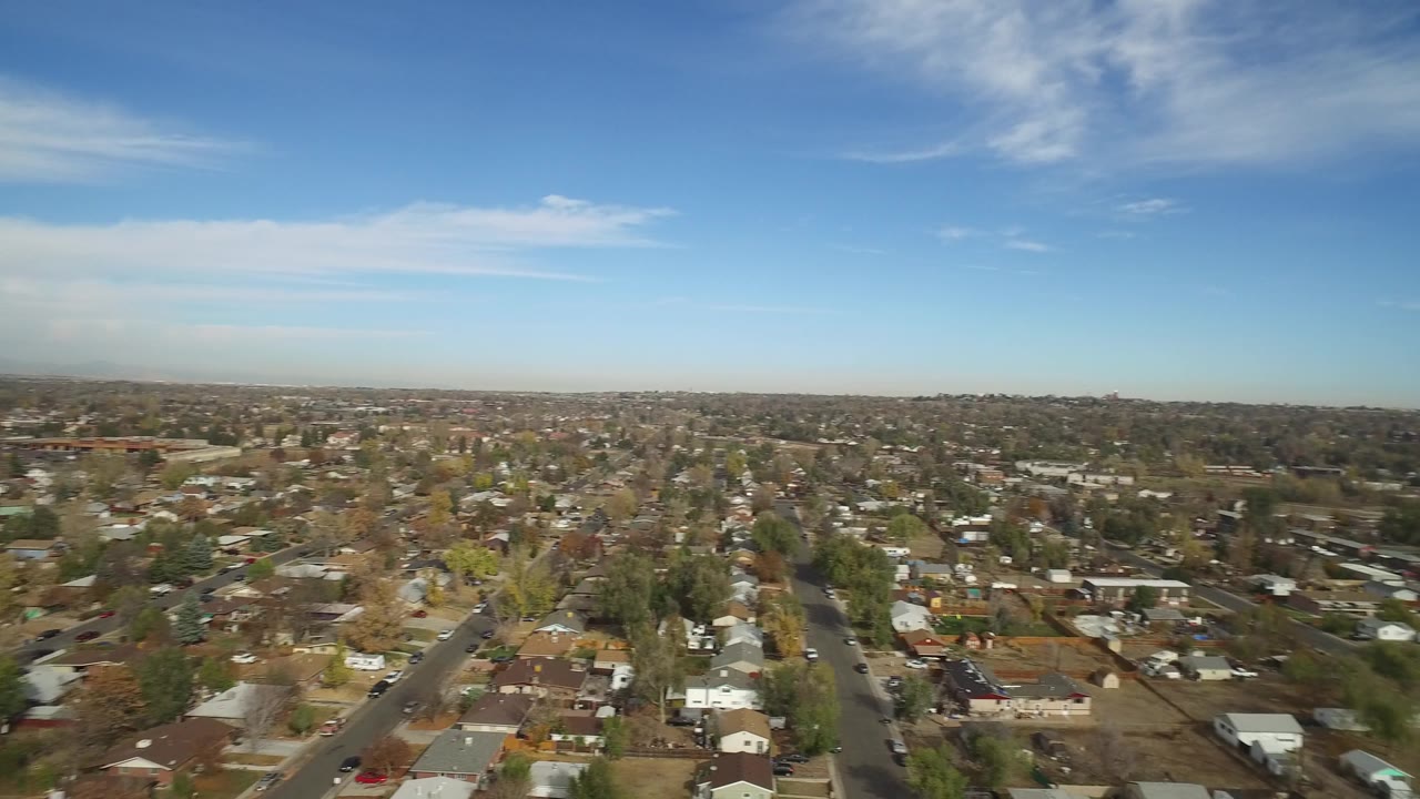 Aerial frame over Hidden Lake, Colorado, with houses and yards in the foreground, straight neighborhood roads through the center, low commercial buildings in the distance, and no visible people.