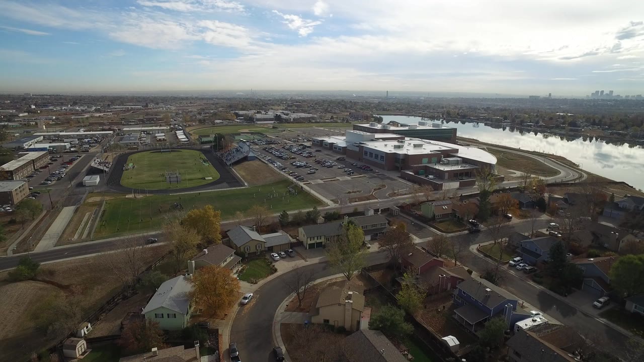 Aerial view of suburban streets, homes, sports field, lake, and distant skyline.
