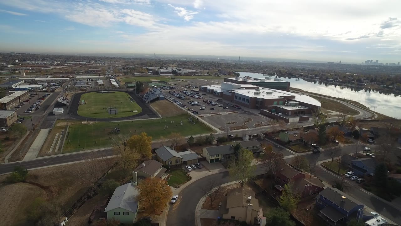 Wide aerial view over Hidden Lake, Colorado, showing green athletic fields, nearby homes, roads, a large white-roofed building, a reflective lake, and no visible people.